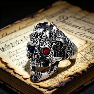 Può includere: Un anello a teschio argentato con occhi di gemme rosse e blu. L'anello presenta dettagli intricati, tra cui teschi più piccoli e motivi ornati. L'anello è esposto su carta vintage con note musicali e testo.