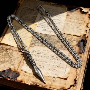 Könnte beinhalten: Eine silberfarbene Anhängerkette mit geflochtener Kette. Der Anhänger ist ein stilisierter Speerkopf mit einem Spiraldetail. Die Kette liegt auf einem offenen antiken Buch mit gealterten Seiten und handschriftlichem Text.