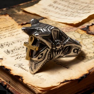 May include: A silver-colored ring with an Egyptian Anubis design, featuring a gold-colored ankh symbol. The ring is detailed with intricate carvings and set against a backdrop of aged paper and a vintage map.