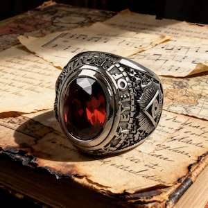 Könnte beinhalten: Ein silberfarbener Ring mit einem ovalen, facettierten, tiefroten Edelstein. Der Ring weist eingravierte Details und Symbole auf, darunter Text um die Lünette und ein Design an der Seite. Der Ring ist vor dem Hintergrund von gealtertem Papier mit handschriftlichem Skript und Noten platziert.