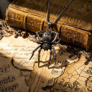May include: A silver spider pendant necklace with a detailed arachnid design. The spider has a textured body and legs, with red accents. The necklace is displayed on aged paper with musical notes and a vintage book in the background.