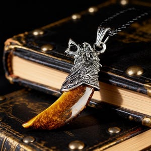 May include: A silver-colored wolf head pendant necklace with a tiger's eye gemstone tooth. The pendant is detailed with intricate carvings and hangs from a black chain. The necklace rests on a stack of antique books.