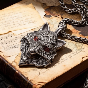 Puede incluir: Un collar con colgante de lobo plateado con ojos de piedras preciosas rojas. El colgante está intrincadamente detallado y descansa sobre un libro abierto con páginas envejecidas y un mapa. También se ve una cadena plateada.