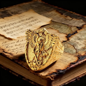 May include: A gold ring with an angel motif, showcasing intricate wing details and a shield-like form. The ring rests on an antique book with aged pages and a map design, giving a vintage appearance.