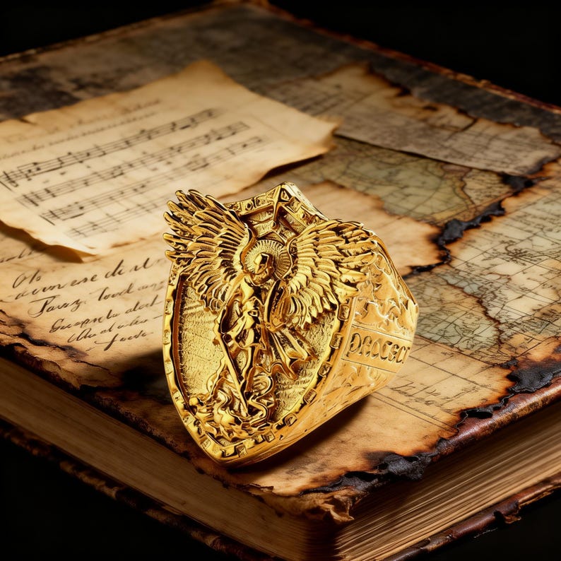 May include: A gold ring with an angel motif, showcasing intricate wing details and a shield-like form. The ring rests on an antique book with aged pages and a map design, giving a vintage appearance.