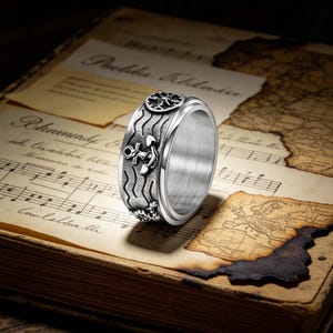 May include: A silver ring with nautical engravings. The ring features a compass design, an anchor, and wave patterns. The ring is set against a backdrop of aged paper with musical notes and a map.