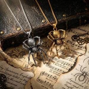 May include: Two spider pendant necklaces. One is silver-toned, the other gold-toned. The spider pendants are detailed with eight legs and a textured body. The necklaces rest on vintage sheet music and a dark leather-bound book.