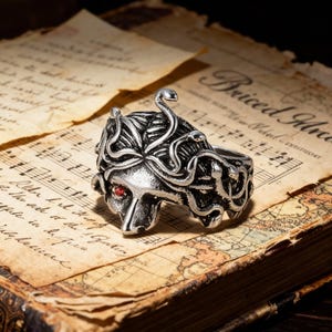 May include: A silver-colored ring featuring a Medusa design with snakes for hair and a red gemstone eye. The ring is set against a backdrop of aged paper with musical notes and text.