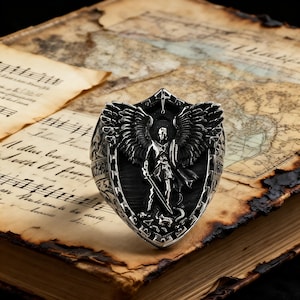May include: A detailed silver ring with a shield design, an angel figure, wings, and a sword. The ring is set against a backdrop of aged paper, sheet music, and a vintage map, creating a historical and artistic aesthetic.