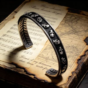 May include: A silver cuff bracelet featuring a series of skull designs. The bracelet has a dark, oxidized finish and is set against a vintage book with musical notes and a map.