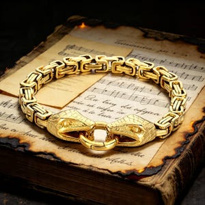 May include: A gold-colored bracelet with a snake head design. The bracelet is made of interlocking links and features two snake heads that connect with a circular clasp. The bracelet is displayed on a vintage book with musical notes.