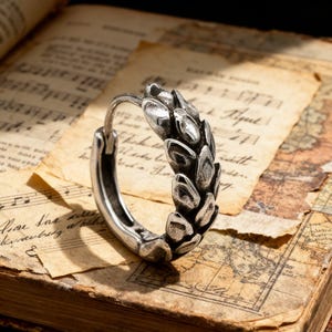 May include: A silver-colored hoop earring with a detailed leaf design. The earring is set against a backdrop of aged paper with musical notes and a map, creating a vintage aesthetic. The earring's design is the focal point.