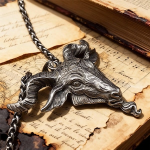 May include: A silver-colored pendant necklace featuring a detailed goat head design. The pendant is intricately sculpted with horns, facial features, and a textured surface. The chain is a braided style. The necklace rests on aged paper with musical notes.