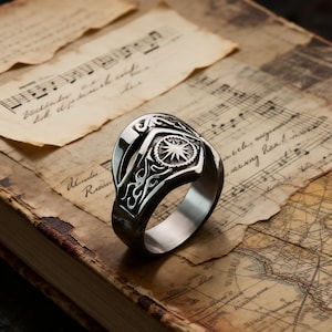 May include: A silver-toned ring with an intricate design featuring a central starburst and flame-like patterns. The ring rests on a vintage book with aged paper and musical notes, creating a classic aesthetic. The ring's design is detailed and eye-catching.