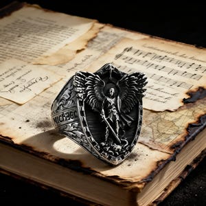 May include: A detailed silver ring featuring a shield design with an angel figure, wings, and the word "MICHAEL". The ring is set against a backdrop of aged paper and musical notes, creating a vintage aesthetic.