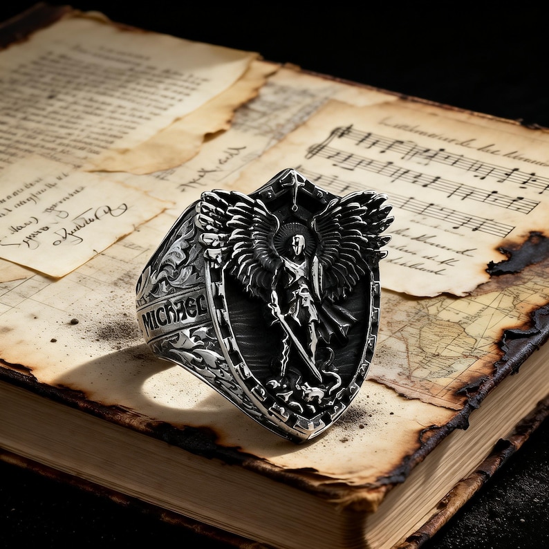 May include: A detailed silver ring featuring a shield design with an angel figure, wings, and the word "MICHAEL". The ring is set against a backdrop of aged paper and musical notes, creating a vintage aesthetic.