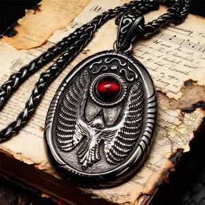 May include: A silver-toned pendant necklace with an oval-shaped pendant featuring a detailed bird design and a red gemstone. The necklace has a braided chain and rests on an open book with aged pages.