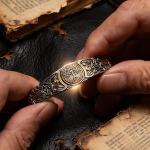 Puede incluir: Brazalete ornamentado de plata y oro con un diseño de árbol de la vida en el centro. El brazalete tiene intrincados nudos celtas y está adornado con pequeñas piedras preciosas verdes. El brazalete está sostenido entre dos manos.