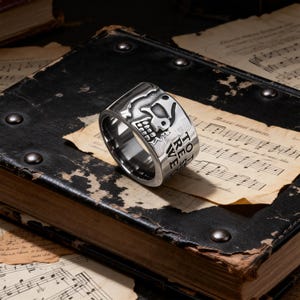 May include: A silver-colored ring with a skull design and the words "TO REST" engraved on the side. The ring is set against a backdrop of aged sheet music and a dark leather-bound book.