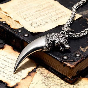 May include: A silver-colored pendant necklace featuring a wolf head and a large claw design. The necklace is on a silver chain. The pendant rests on a vintage book with aged paper and a map.