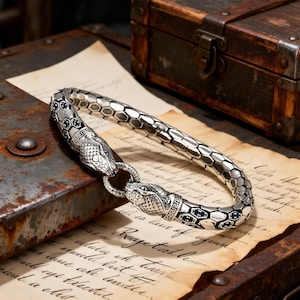 May include: A silver bracelet with a snake design. The bracelet features two snake heads connected by a ring clasp. The bracelet has a hexagonal pattern and is decorated with blue accents. The bracelet is resting on a vintage paper.