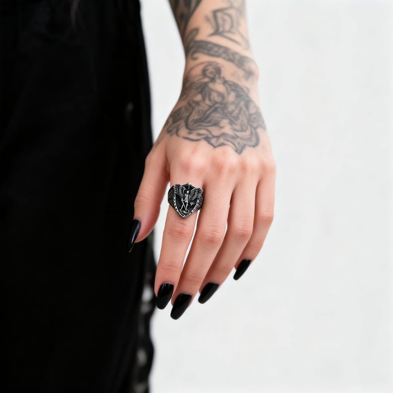 May include: A black shield-shaped ring with a detailed design featuring wings and a central figure. The ring is worn on a hand with black nail polish, set against a white background. The ring's design is intricate and gothic.