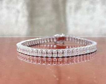 Op de afbeelding: Een zilverkleurige tennisarmband met een doorlopende rij rechthoekige, baguette-geslepen heldere stenen. De armband heeft een metallic afwerking en een zichtbaar sluitmechanisme aan de achterkant dat licht op het oppervlak weerkaatst.