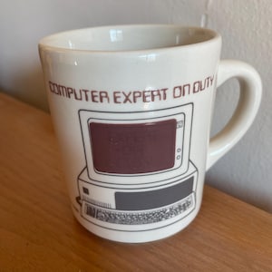 Pode incluir: Caneca de cerâmica creme com uma alça. A caneca apresenta um gráfico de computador retrô com uma tela bordô e o texto "COMPUTER EXPERT ON DUTY" em letras bordô. A caneca está em uma superfície de madeira.