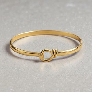 Może przedstawiać: Złota bransoletka typu bangle z unikalnym wzorem węzła. Bransoletka wykonana jest z błyszczącego, gładkiego metalu i posiada okrągłe zapięcie. Całość jest prosta, a zarazem elegancka, odpowiednia na co dzień lub specjalne okazje.