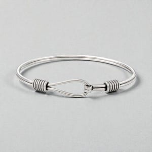 Może przedstawiać: Srebrna bransoletka typu obręcz z zapięciem na haczyk. Bransoletka ma prosty, elegancki design z zaokrągloną opaską i dekoracyjnymi, zwiniętymi akcentami w pobliżu zapięcia. Zapięcie to unikalne, bezpieczne zamknięcie.