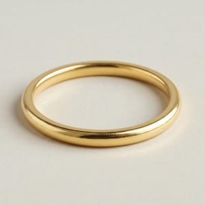 Puede incluir: Un anillo dorado con una banda lisa y redondeada. El anillo tiene un acabado pulido, que refleja la luz. La joya se encuentra sobre un fondo blanco liso.