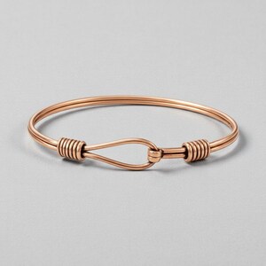 May include: A copper-coloured bangle bracelet with a hook and eye closure. The bracelet features a simple, elegant design with wrapped wire detailing at the clasp. The bracelet is shown on a light grey background.