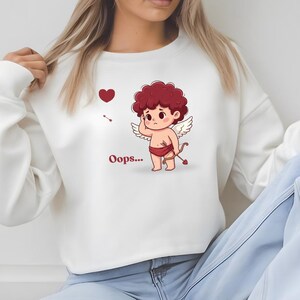 May include: White sweatshirt featuring a cartoon Cupid with red curly hair, wings, and a bow and arrow. A red heart and the word "Oops..." are also visible. The sweatshirt is a casual style.