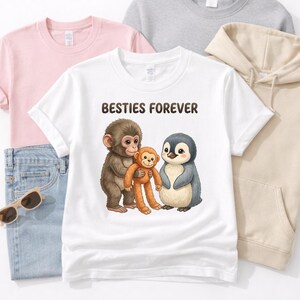 May include: White t-shirt with the text "BESTIES FOREVER" and a graphic of a monkey, a smaller orange monkey, and a penguin. Other shirts in pink and gray are visible, along with a pair of blue jeans and sunglasses.