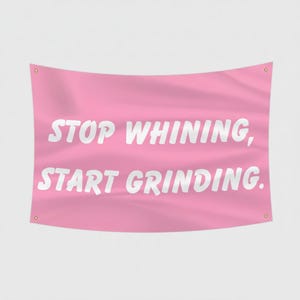 Puede incluir: Bandera rosa con texto blanco "STOP WHINING, START GRINDING." La bandera tiene ojales en cada esquina para colgar. El fondo es de color rosa claro liso.