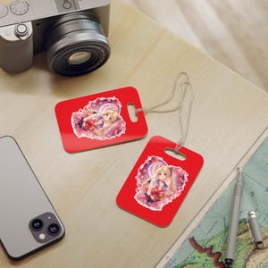 May include: Two red luggage tags with a cartoon image of a blonde girl in a sailor uniform. The tags have a white string attached and are on a light-colored surface. A camera and a smartphone are also visible.