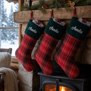Puede incluir: Tres calcetines navideños con un patrón de cuadros rojos y verdes y un puño de terciopelo verde oscuro. Cada calcetín está bordado con el nombre Aurelia en letra blanca. Los calcetines cuelgan de lazos rojos en una repisa de piedra.