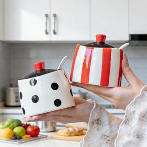 Può includere: Due zuccheriere in ceramica, una con pois neri e l'altra con strisce rosse e bianche. Ogni ciotola ha un coperchio nero con un pomello rosso e un cucchiaino bianco. Le ciotole sono tenute davanti a uno sfondo di cucina.