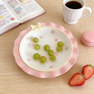 May include: A decorative pink and white ceramic plate with a scalloped edge and a bow. The plate holds green grapes and is decorated with pink polka dots. A cup of coffee, a pink macaron, and two strawberry halves are also present.