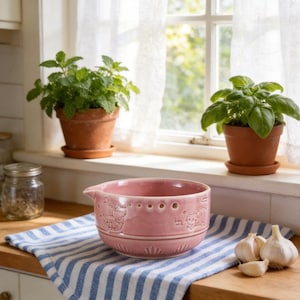 Peut inclure: Un saladier en céramique rose avec des motifs d'animaux en relief et un bec verseur est posé sur un torchon à rayures bleues et blanches. Deux herbes en pot et des gousses d'ail sont également visibles.