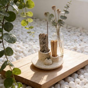 Może przedstawiać: Beżowy kamienny uchwyt na kadzidło z wiązką szałwii, patyczkiem do kadzidła i patyczkami palo santo. Uchwyt stoi na drewnianej tacy z małym wazonem z suszonymi kwiatami. Zielone liście i białe kamienie w tle.