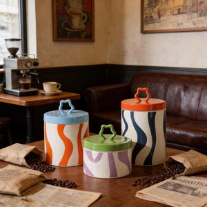 Puede incluir: Juego de tres botes de cerámica con tapas. Los botes tienen una base blanca moteada con diseños ondulados coloridos en naranja, azul marino y morado. Cada tapa es de un color diferente: azul claro, naranja y verde. Asas en forma de estrella.