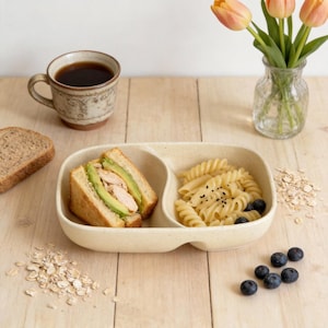 May include: A beige divided plate holds a sandwich with avocado and chicken, and pasta with blueberries. A mug of coffee, a slice of bread, and a vase of tulips are also on the wooden surface. Oatmeal is scattered around the plate.