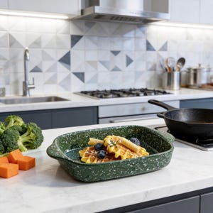 May include: A green speckled rectangular baking dish with handles, filled with waffles, blueberries, and a rolled pastry. Broccoli and orange cubes are on the counter. A black skillet sits on the stove.