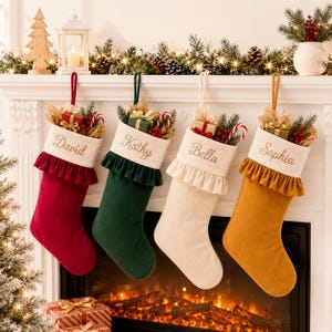 Puede incluir: Cuatro calcetines navideños de terciopelo en rojo, verde oscuro, crema y amarillo mostaza, cada uno con un puño blanco bordado con los nombres David, Kathy, Bella y Sophia, decorados con cajitas de regalo y ramas de pino.