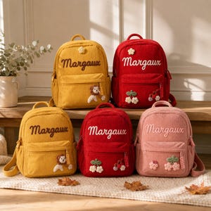Puede incluir: Cinco mochilas de pana en amarillo, rojo y rosa, cada una con el nombre bordado Margaux. Las bolsas tienen un bolsillo frontal con cremallera, correas ajustables y aplicaciones decorativas que incluyen un león, cerezas y flores.