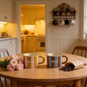 May include: Two ceramic mugs, one with "MAMA" and the other with "PAPÁ", sit on a wooden table. The "MAMA" mug has pink and yellow stripes, the "PAPÁ" mug has blue and brown stripes. A bouquet of pink flowers and a small bag of coffee beans are also on the table.