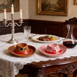 May include: Two ceramic cowboy hat-shaped dishes, one brown with sunglasses and the other pink with cherries, are on a lace tablecloth. A silver candelabra with lit candles, a wine decanter, and a plate of food are also on the table.