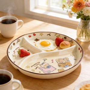 May include: A cream-coloured divided ceramic plate with a cartoon design, holding a fried egg, strawberries, and a small roll. The plate has brown trim and is decorated with hearts, stars, and the text "Shopping Time".