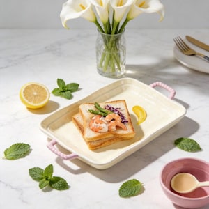 Può includere: Un piatto da portata rettangolare bianco sporco con manici rosa contiene due fette di pane tostato farcite con gamberi, asparagi e cavolo rosso. Sono presenti anche mezzo limone, foglie di menta e una piccola ciotola rosa con un cucchiaio.